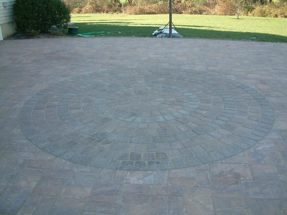 A stone patio featuring a circular pattern made of grey pavers, surrounded by a light brown brick walkway and a green lawn.