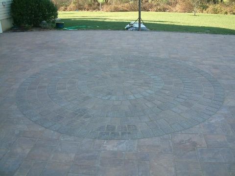 A stone patio featuring a circular pattern made of grey pavers, surrounded by a light brown brick walkway and a green lawn. A stone patio featuring a circular pattern made of grey pavers, surrounded by a light brown brick walkway and a green lawn.