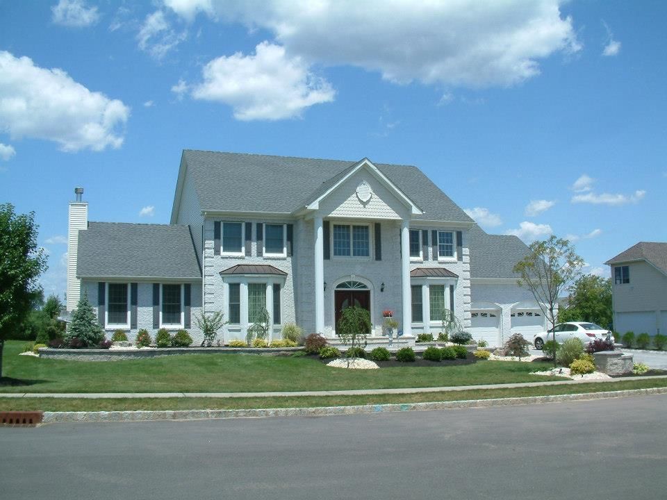 A two-story light gray house with a prominent pillared porch, dark shutters, and a lawn under a blue, cloudy sky.