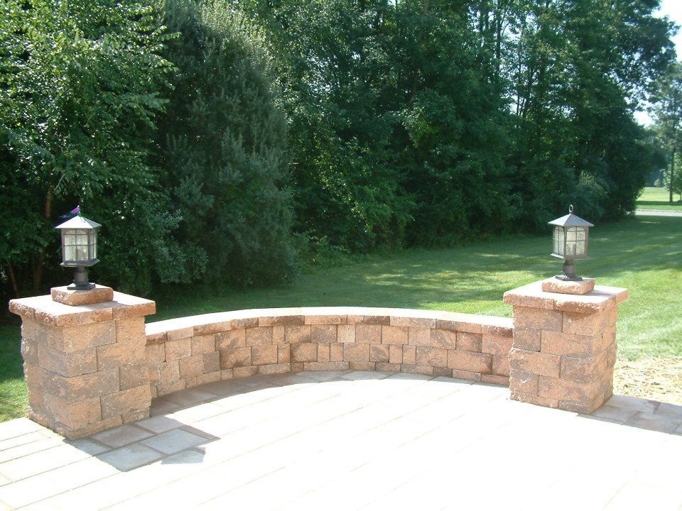 A curved, tan stone seating wall with a lantern mounted on a pillar at each end, set on a patio overlooking a lawn.