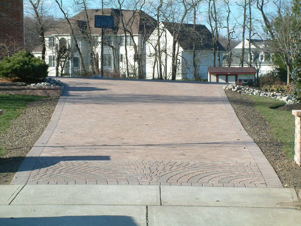 A paved driveway with a decorative fan-pattern border leads toward a two-story suburban house on a sunny day.