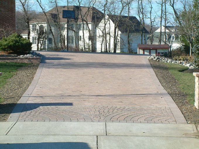 A paved driveway with a decorative fan-pattern border leads toward a two-story suburban house on a sunny day. A paved driveway with a decorative fan-pattern border leads toward a two-story suburban house on a sunny day.