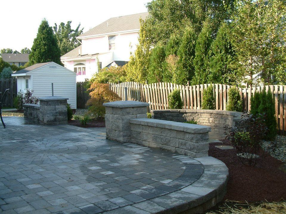 A paved backyard patio featuring stone retaining walls, pillars, and landscaping, set against a wooden fence and house.