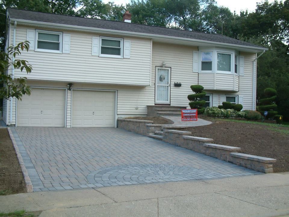 A beige split-level house featuring a paver driveway, stone retaining walls, and a new paved walkway to the front door. A beige split-level house featuring a paver driveway, stone retaining walls, and a new paved walkway to the front door.