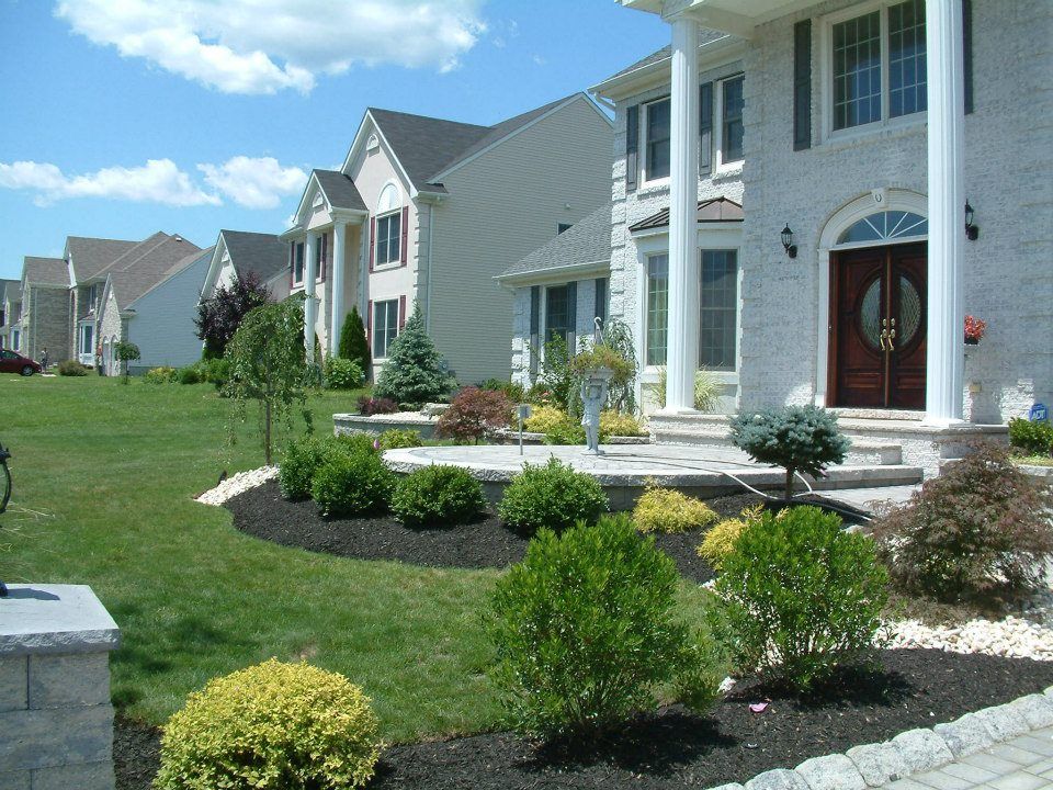 A row of suburban homes with landscaped front lawns, dark mulch beds, and a stone porch on a sunny, blue-sky day.