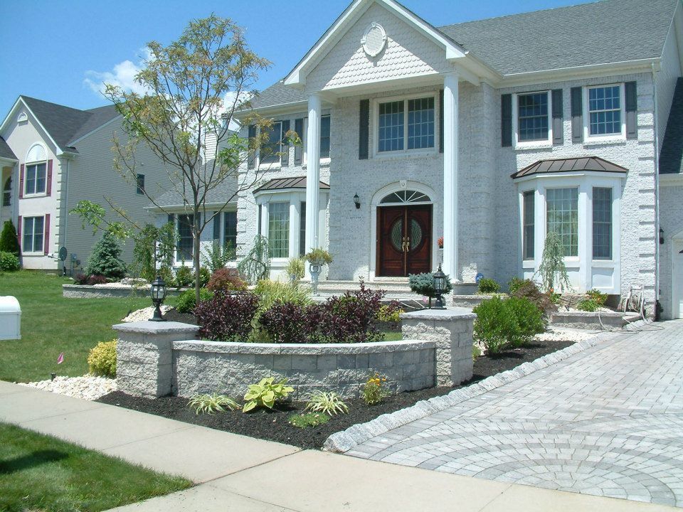 A two-story white house with a stone retaining wall, front garden, and paved driveway on a sunny day.