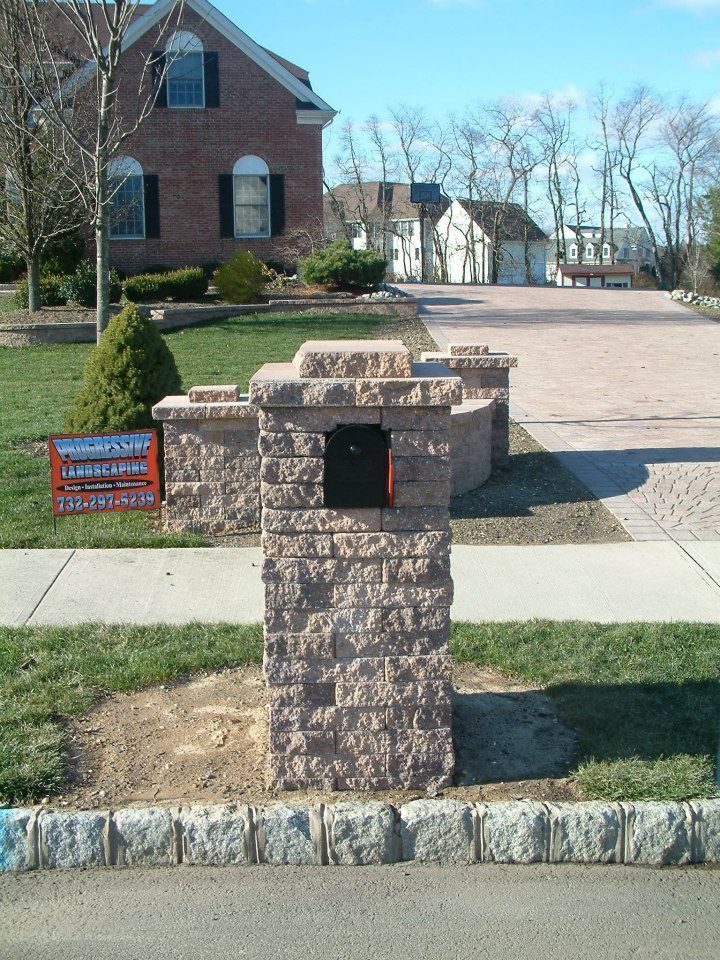 A tan, block-style stone mailbox column stands on a residential lawn in front of a brick house.
