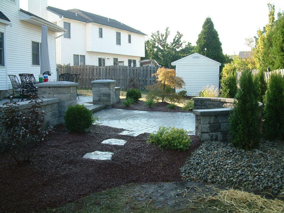 A backyard patio with stone pavers, a small retaining wall, dark mulch, stone steps, and landscaping in a suburban yard.