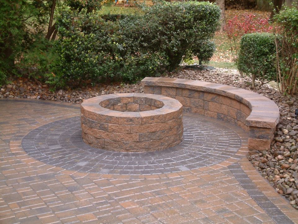 A circular stone fire pit sits on a patterned brick patio with a curved stone bench in a landscaped garden.