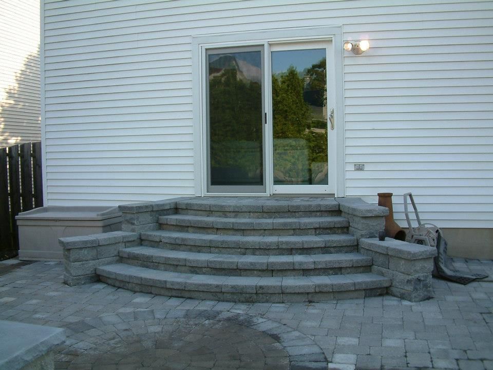 A set of three curved gray stone steps leading up to a sliding glass door on a white-sided house with a paved patio.
