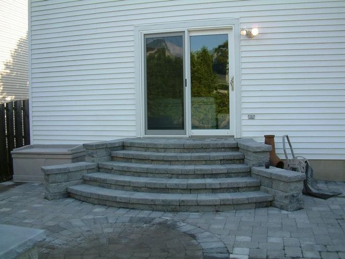 A set of three curved gray stone steps leading up to a sliding glass door on a white-sided house with a paved patio. A set of three curved gray stone steps leading up to a sliding glass door on a white-sided house with a paved patio.