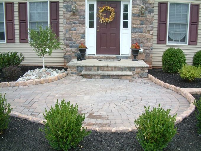 A circular stone patio leads to a stone porch and burgundy front door with a wreath, surrounded by bushes and dark mulch. A circular stone patio leads to a stone porch and burgundy front door with a wreath, surrounded by bushes and dark mulch.