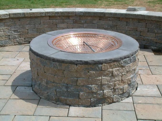 A circular stone fire pit with a copper basin sits on a paved patio in front of a matching curved stone retaining wall. A circular stone fire pit with a copper basin sits on a paved patio in front of a matching curved stone retaining wall.