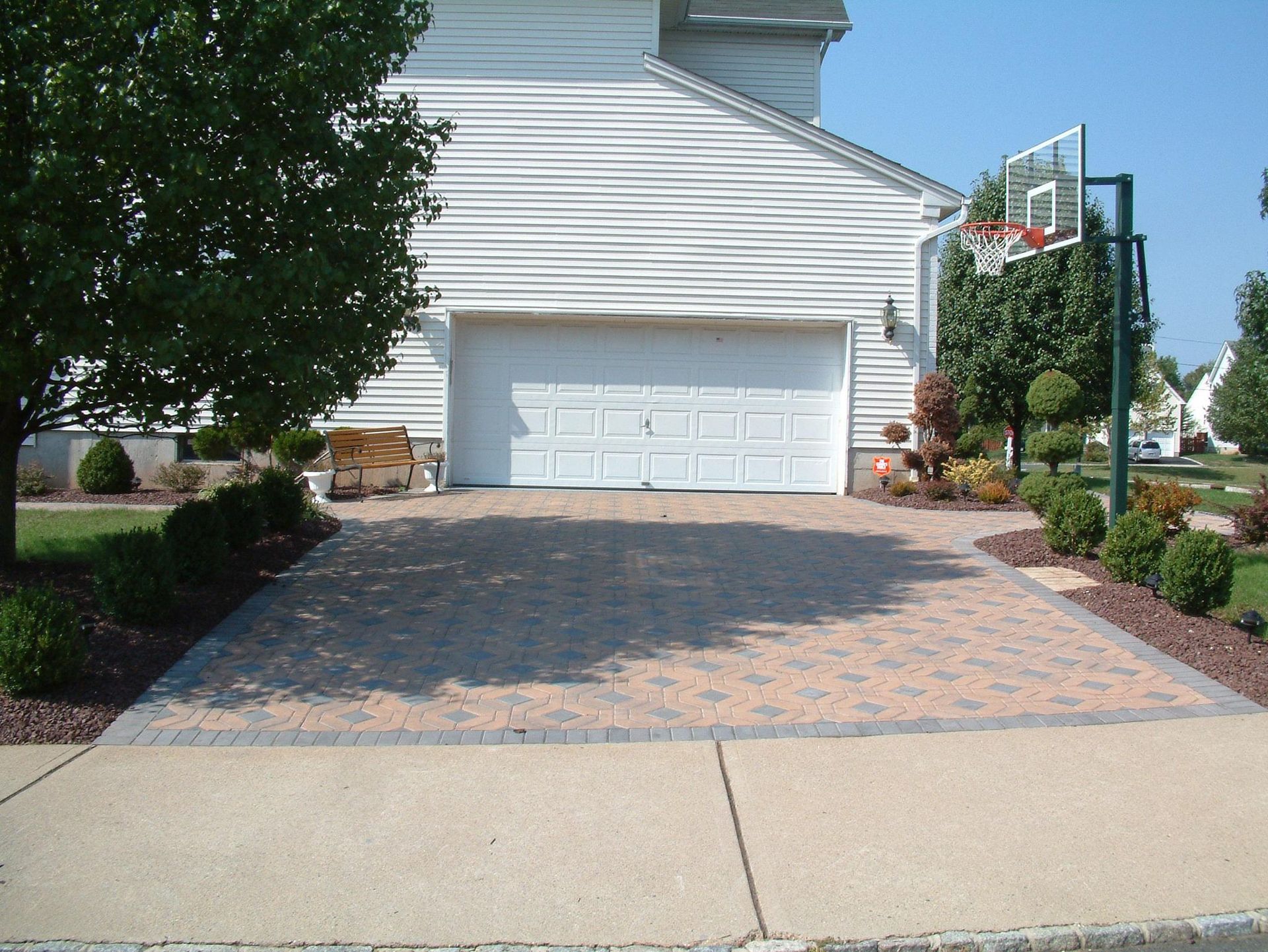A tan paver driveway leading to a white two-car garage, with a tree, manicured shrubs, and a basketball hoop to the side.