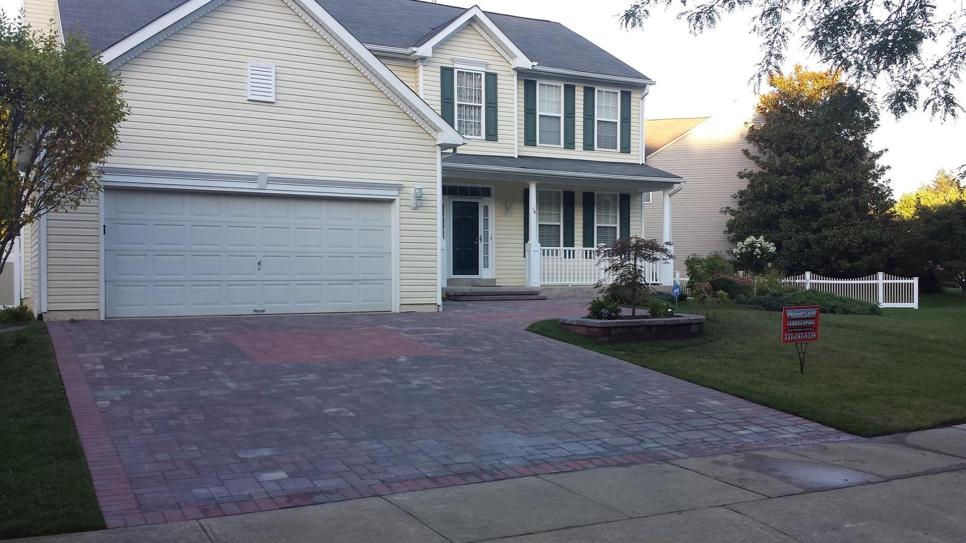 A two-story yellow house with a gray shingled roof, a paved driveway, and a white picket fence in the front yard.