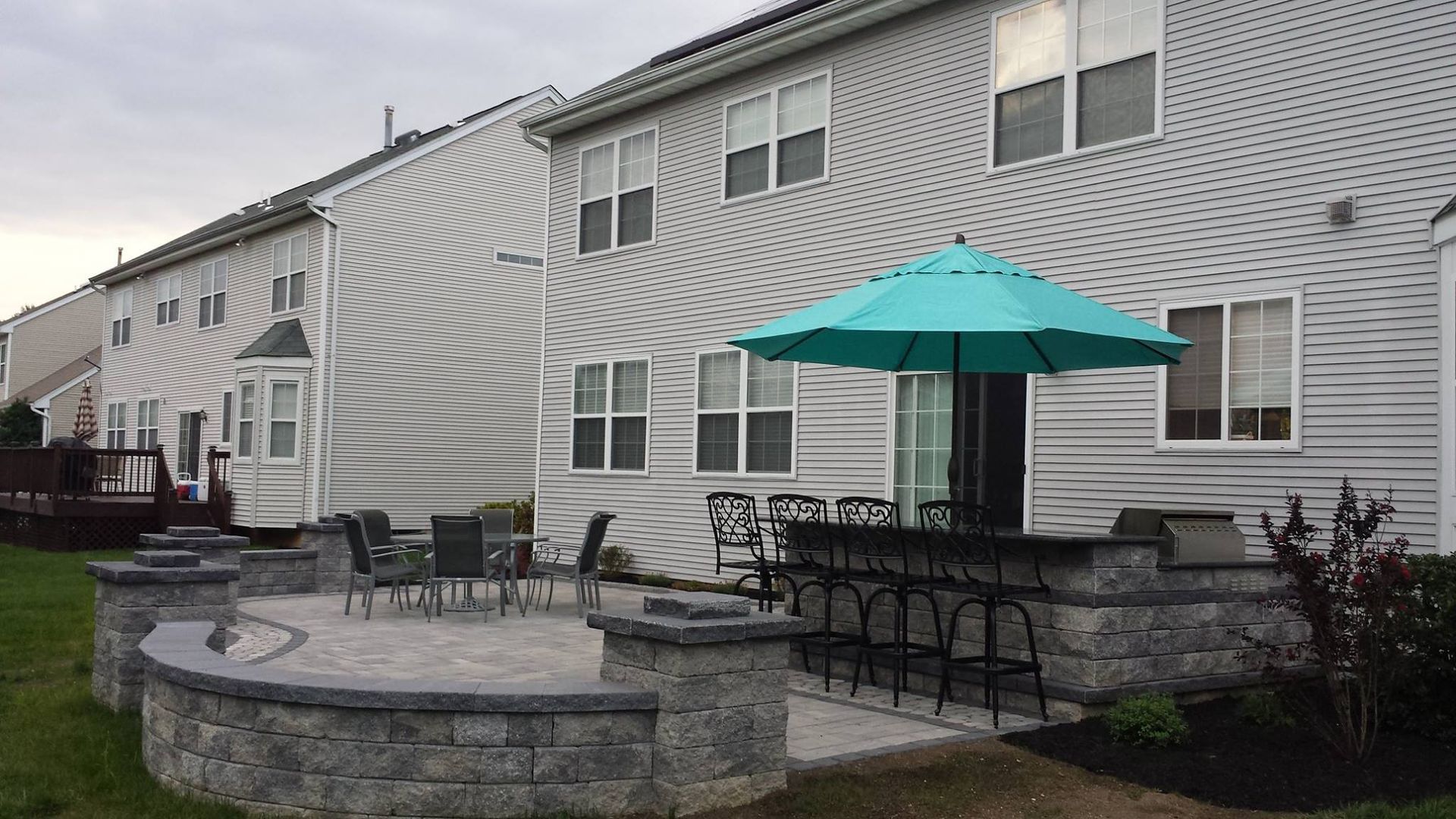 A multi-level stone patio with a built-in stone bar, high chairs, an umbrella, and a small dining set behind a suburban home.