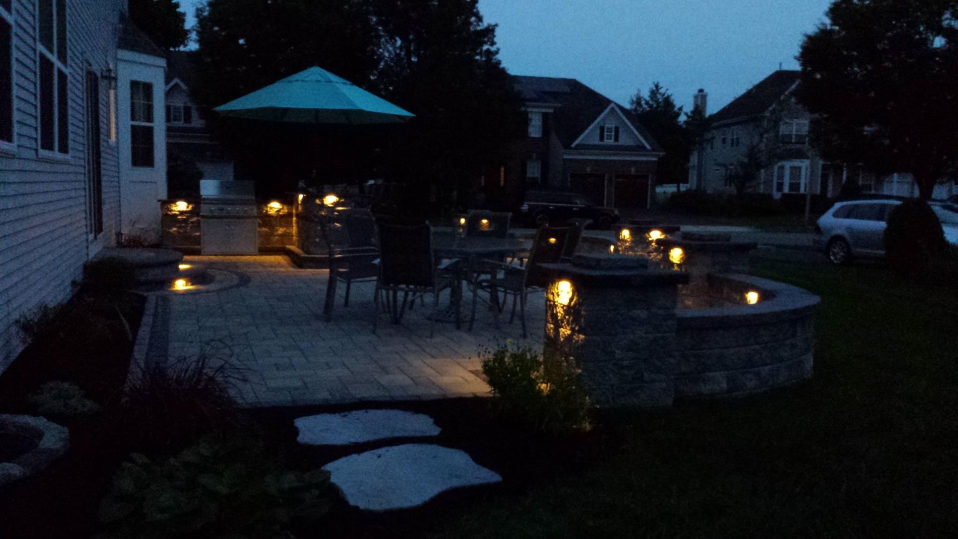 An illuminated stone patio with outdoor furniture, a grill, and an umbrella at dusk, viewed from a backyard.