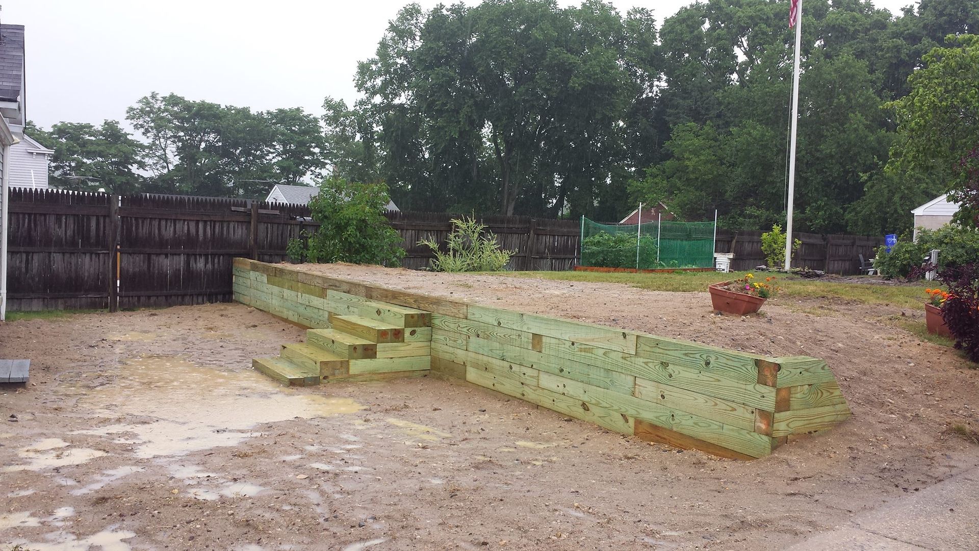 A low, light-green timber retaining wall with steps built into a sloped backyard area with dirt ground and a wooden fence.