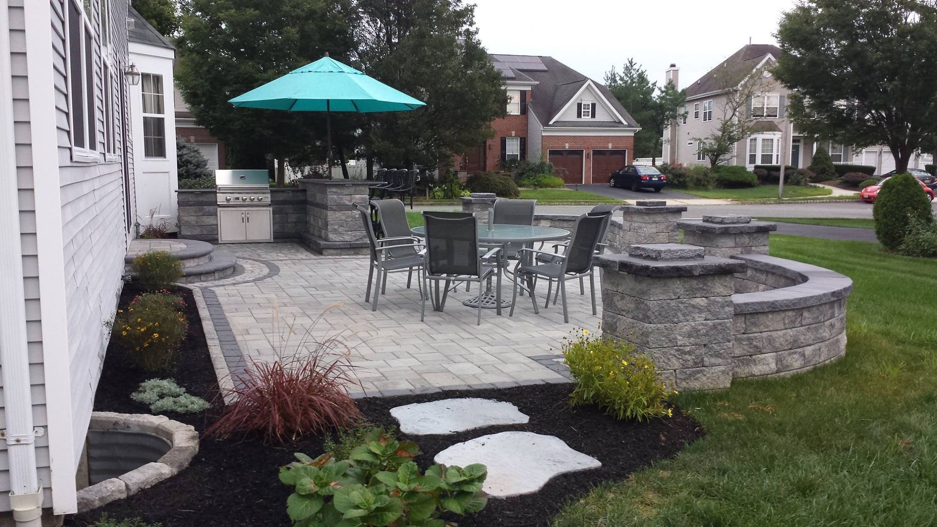 A stone paver patio featuring a dining table, chairs, a turquoise umbrella, and a built-in outdoor grill area.