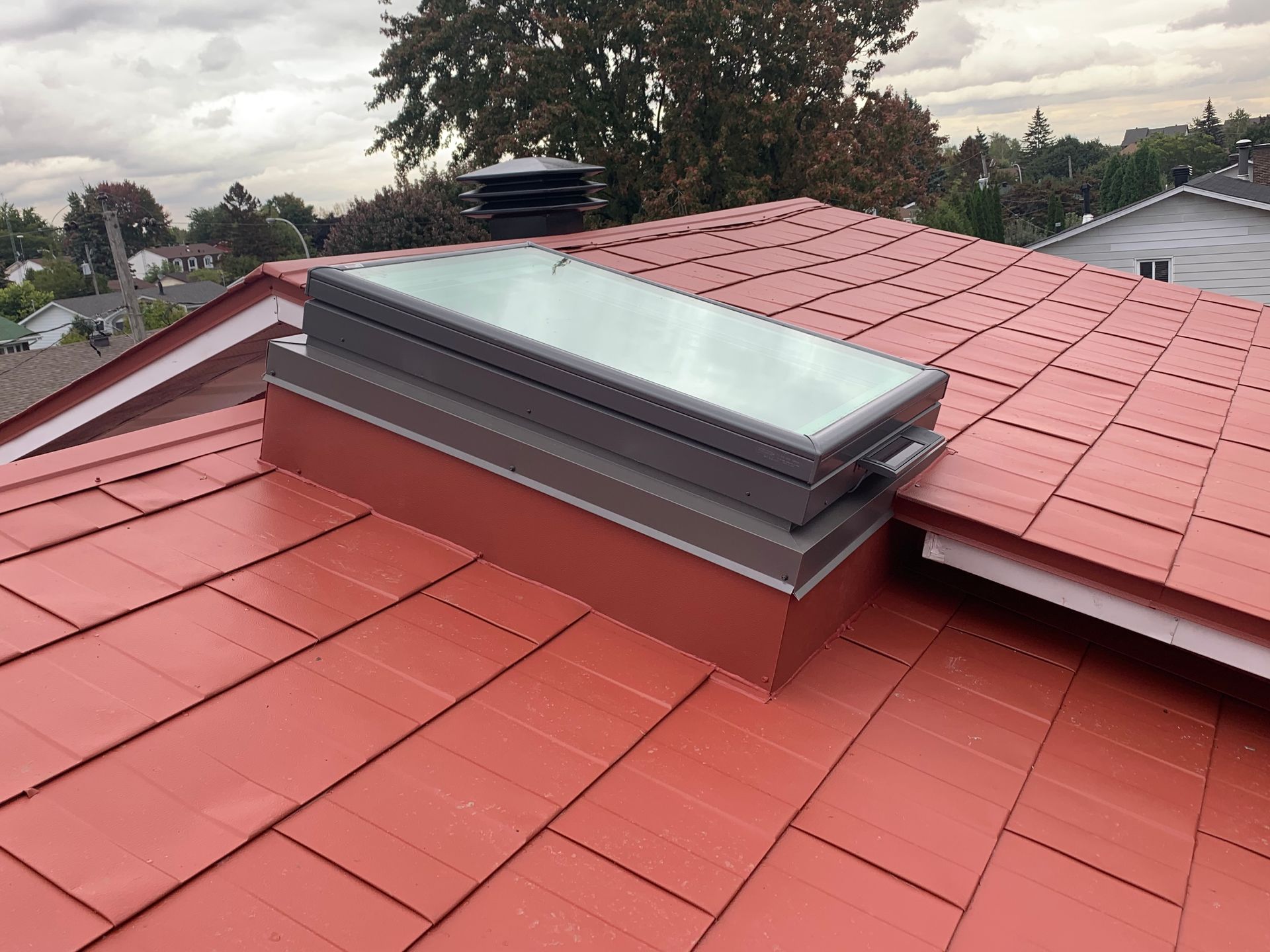 Toit de tuiles rouges avec une lucarne rectangulaire à cadre argenté. Ciel couvert en arrière-plan.