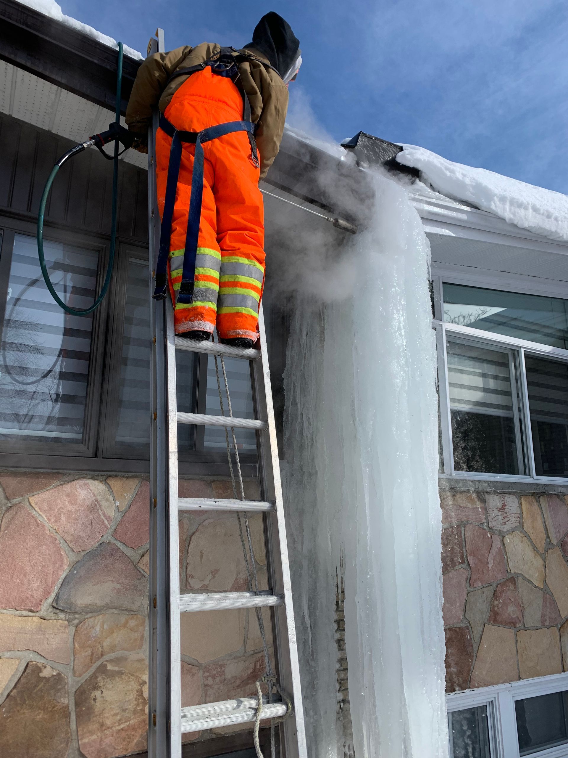 A man is standing on a ladder cleaning icicles on the side of a building.