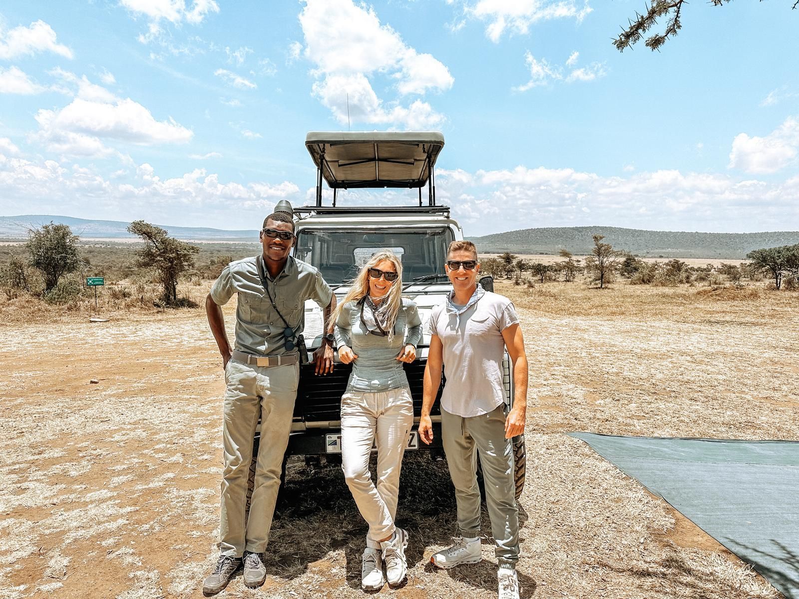 Three people standing beside an off-road vehicle in a dry savanna landscape under a blue sky.