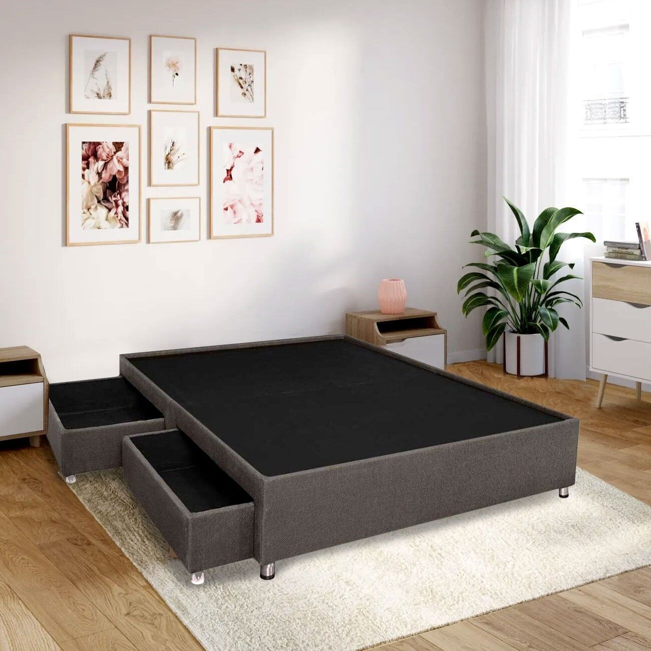 Estructura de cama gris oscuro con cajones, sobre una alfombra, en un dormitorio con obras de arte, una planta y mesitas de noche.