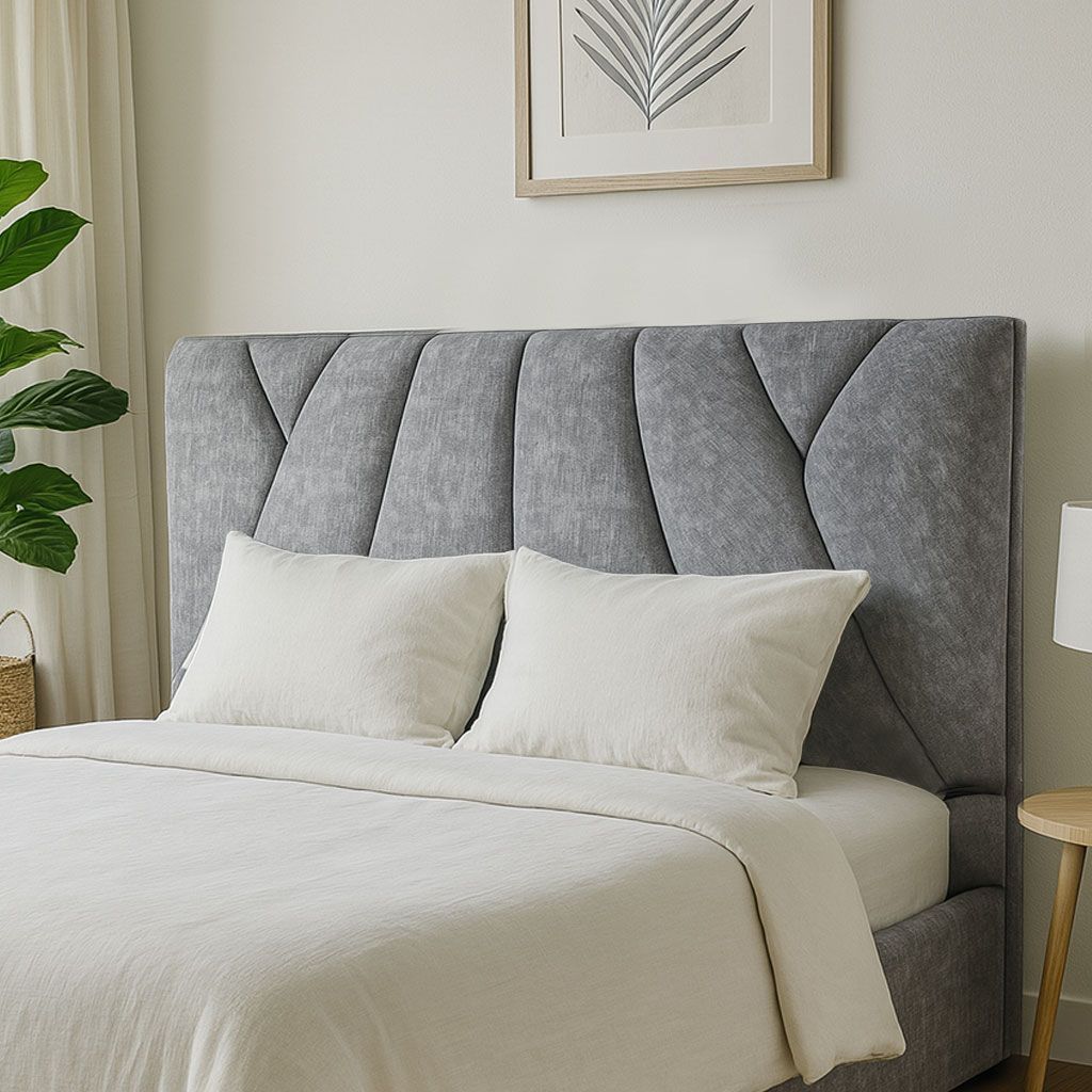 Cama tapizada en gris con ropa de cama y almohadas blancas, contra una pared clara con arte enmarcado.