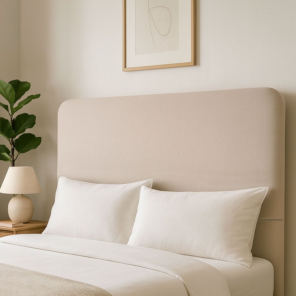 Un cabecero tapizado en color beige con dos almohadas blancas y ropa de cama, junto a una lámpara y una planta en maceta.