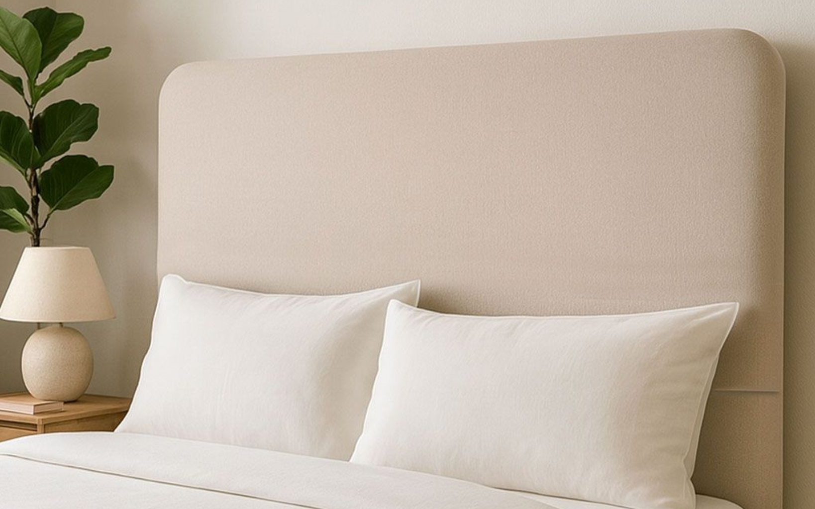 Un cabecero tapizado de color beige detrás de dos almohadas blancas sobre una cama, junto a una pequeña lámpara y una planta en maceta.