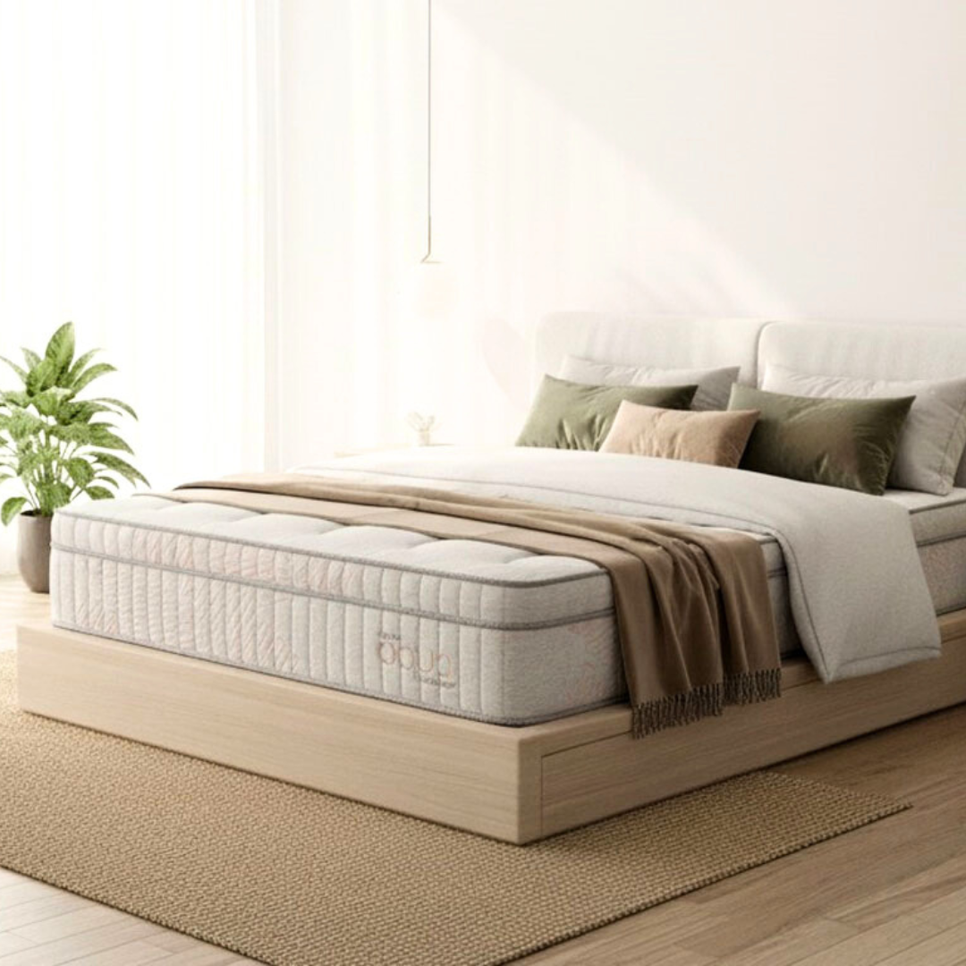 Un colchón de textura blanca reposa sobre una estructura de cama de madera clara en una habitación soleada con una planta y almohadas.