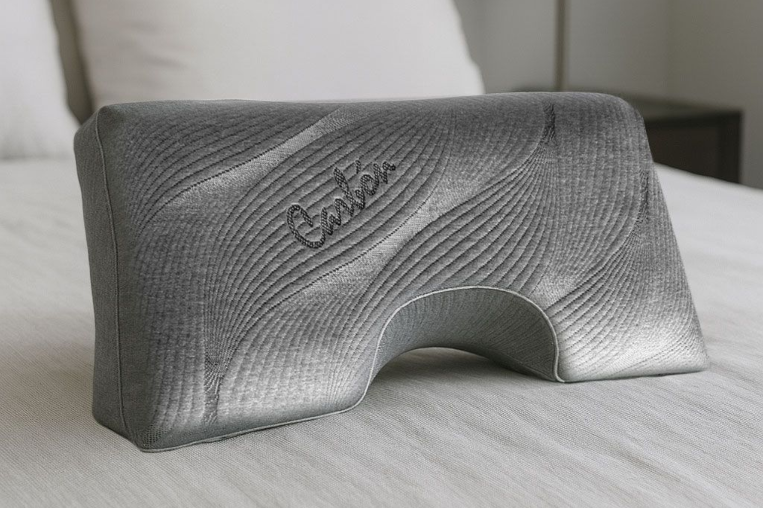Almohada de rodilla de color gris sobre una cama con un recorte curvo y diseño en relieve.