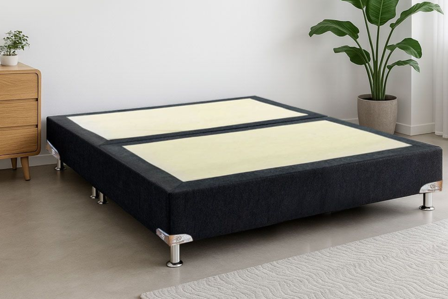 Una base de colchón tapizada en gris oscuro con patas plateadas sobre un suelo de hormigón, junto a una cómoda de madera y una planta en maceta.