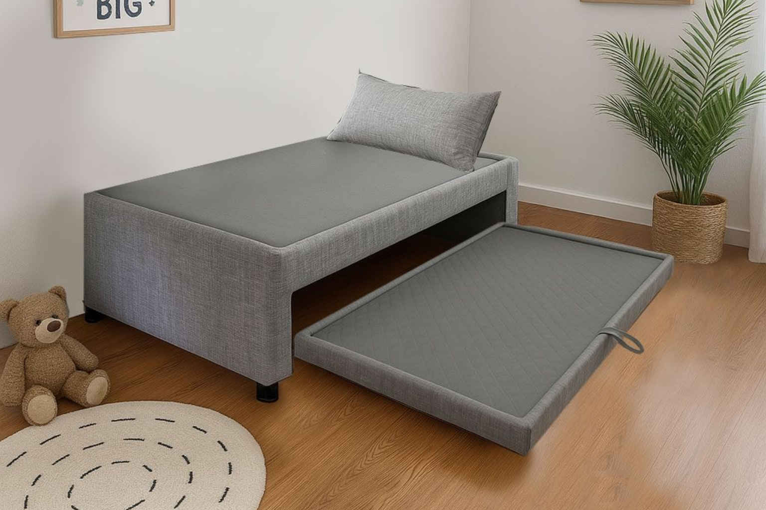 Una cama nido tapizada en gris en un dormitorio, con la sección extraíble extendida junto a un osito de peluche y una alfombra redonda.