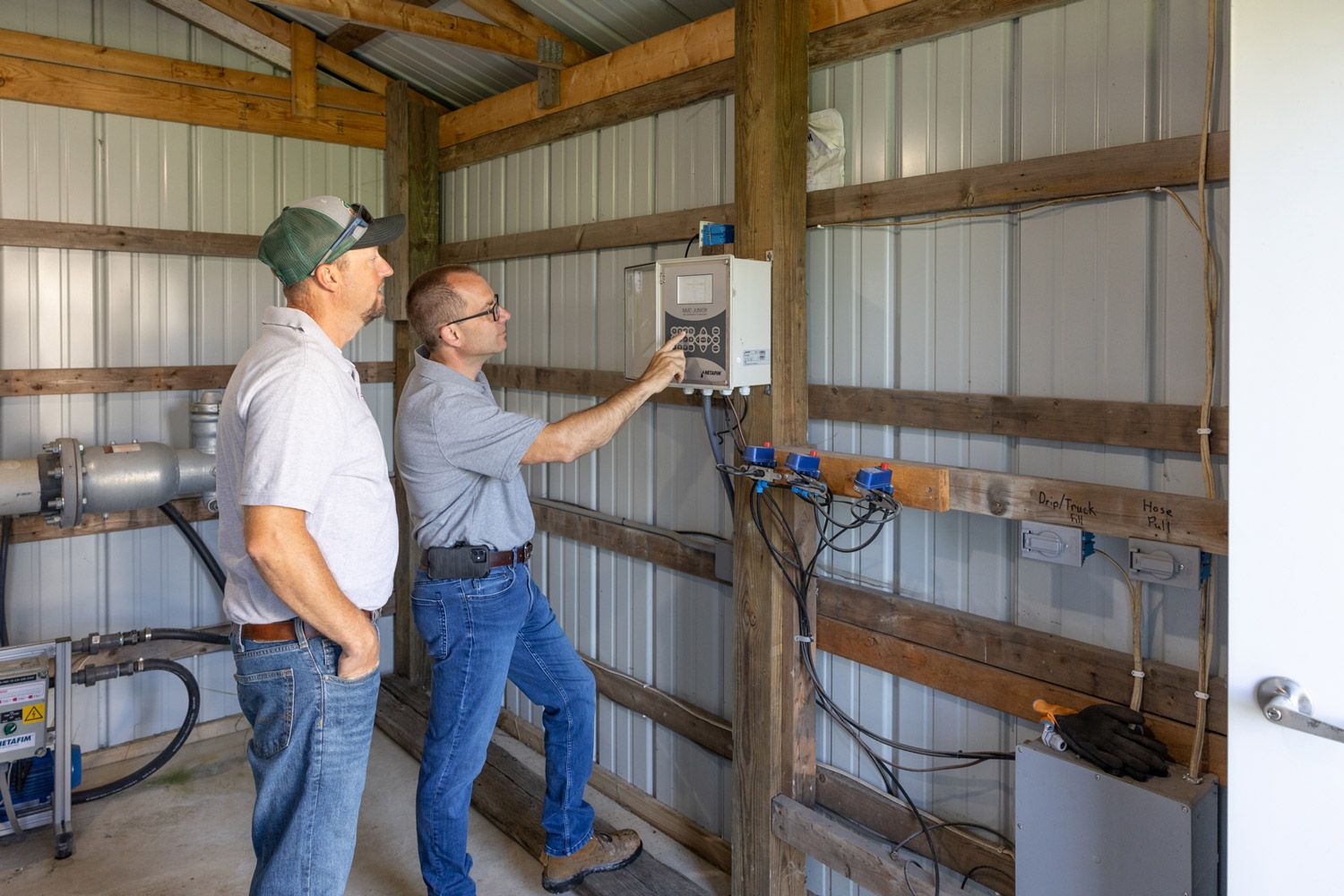 Two men are standing in a barn looking at subsurface irrigation controls.