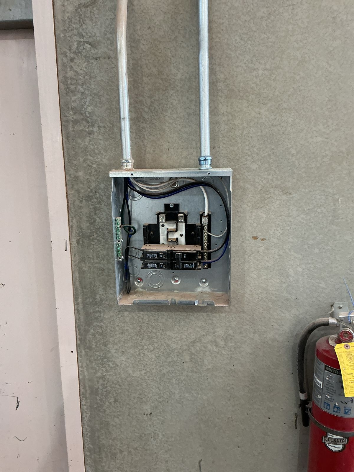 Electrical panel box on a concrete wall with conduit, next to a red fire extinguisher.
