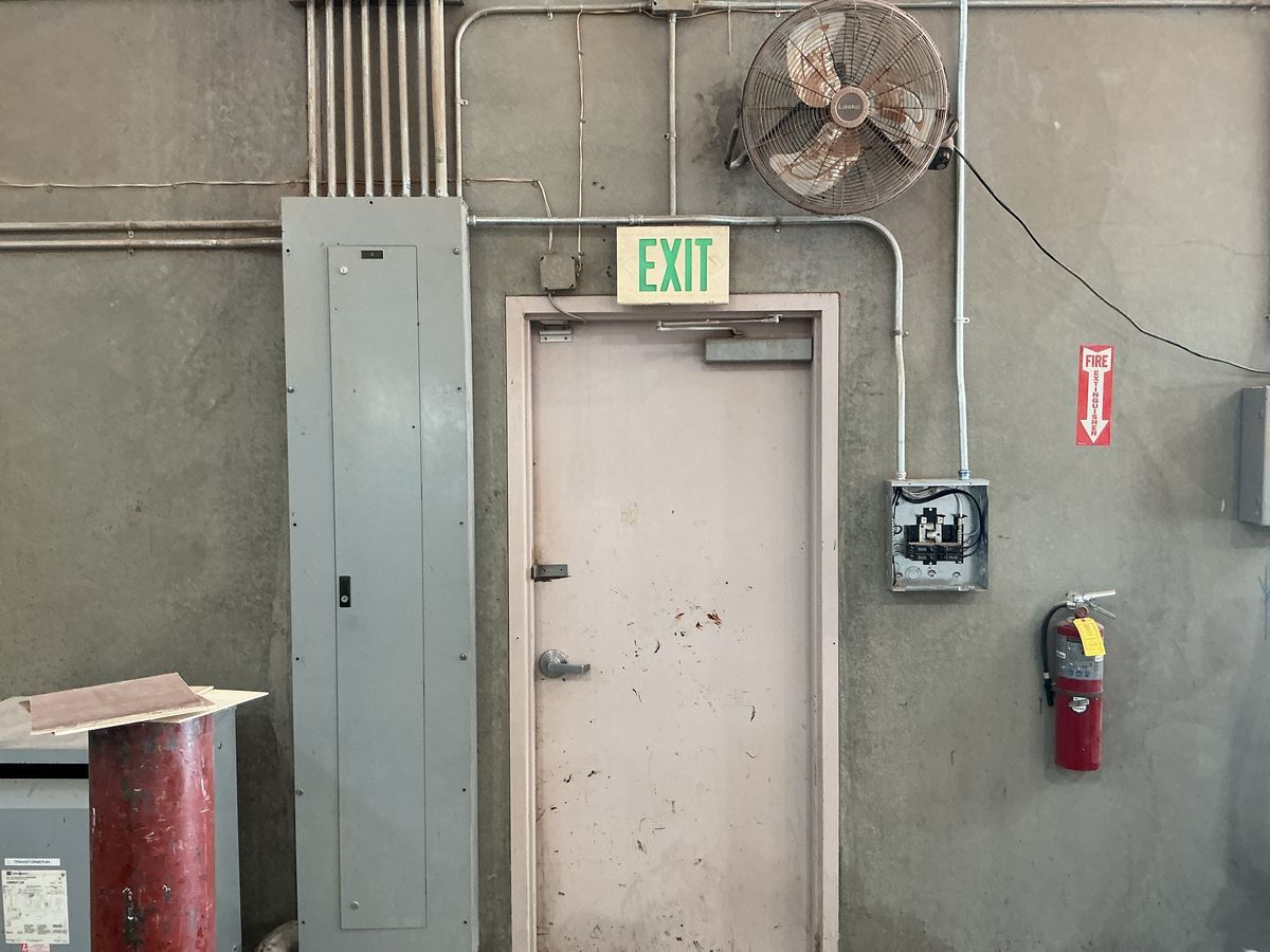 Industrial room interior with an exit door, electrical panel, fan, and fire extinguisher.