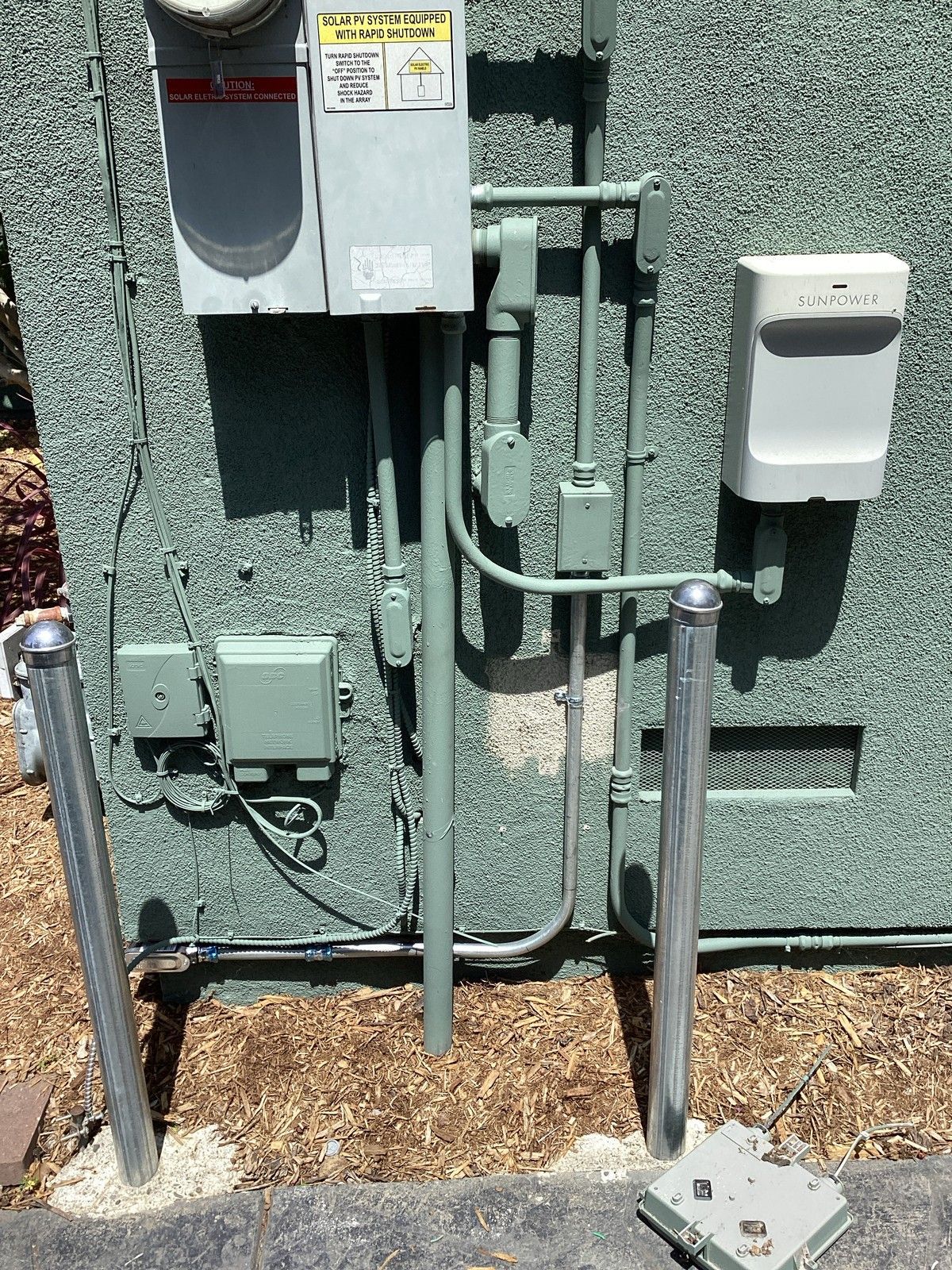Green utility box with electrical conduit and two metal bollards protecting it.