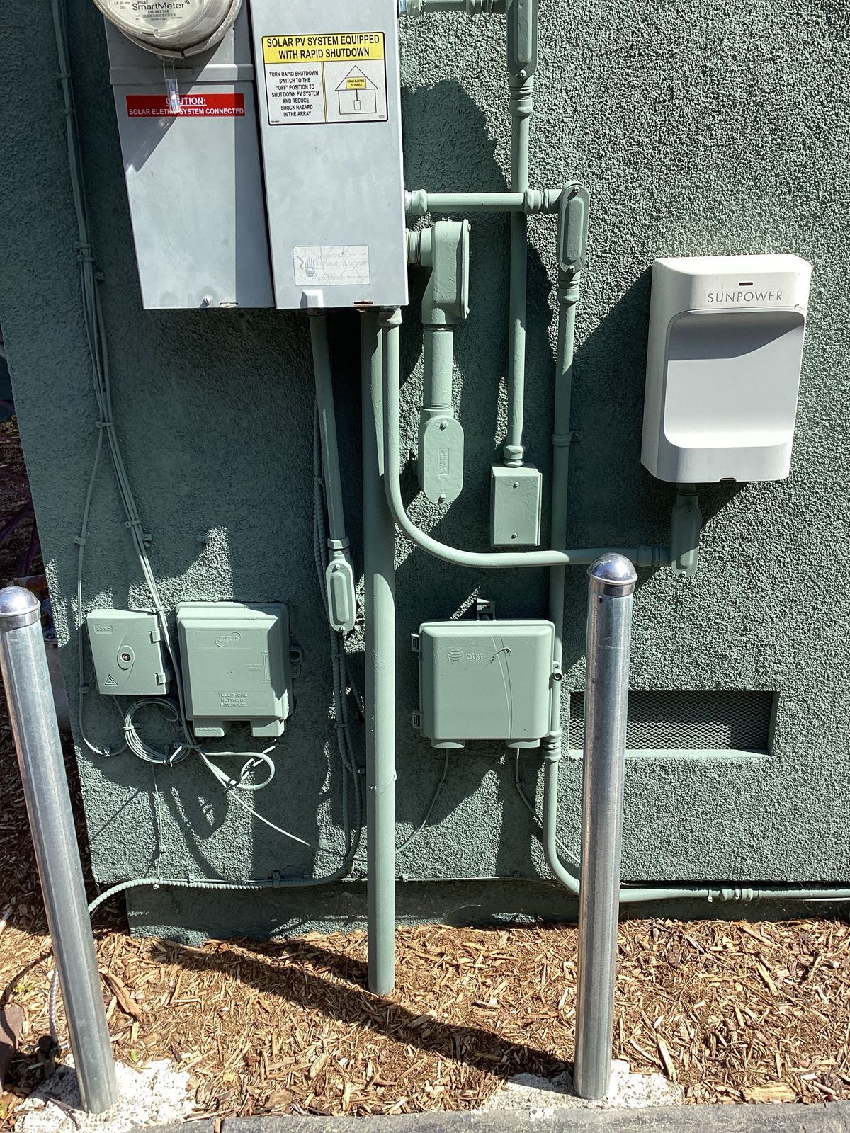 Electrical boxes and conduit on a textured, light gray wall, with two silver bollards at the base.