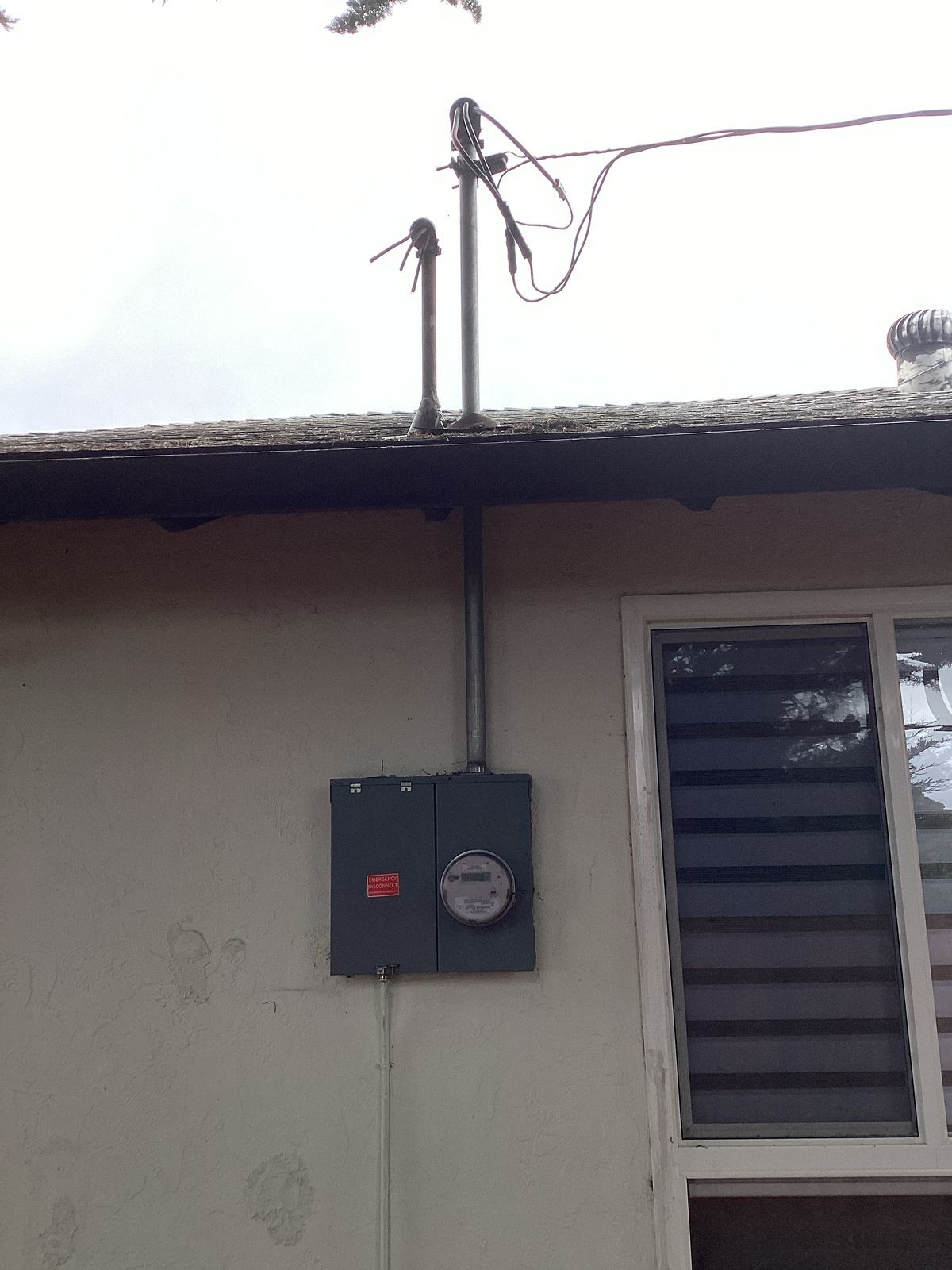 Gray electrical meter box mounted on a light beige house wall with a window. Electrical wires run above.