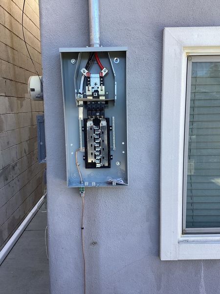 Open electrical panel mounted on a stucco wall next to a window; conduit above, wires visible inside.
