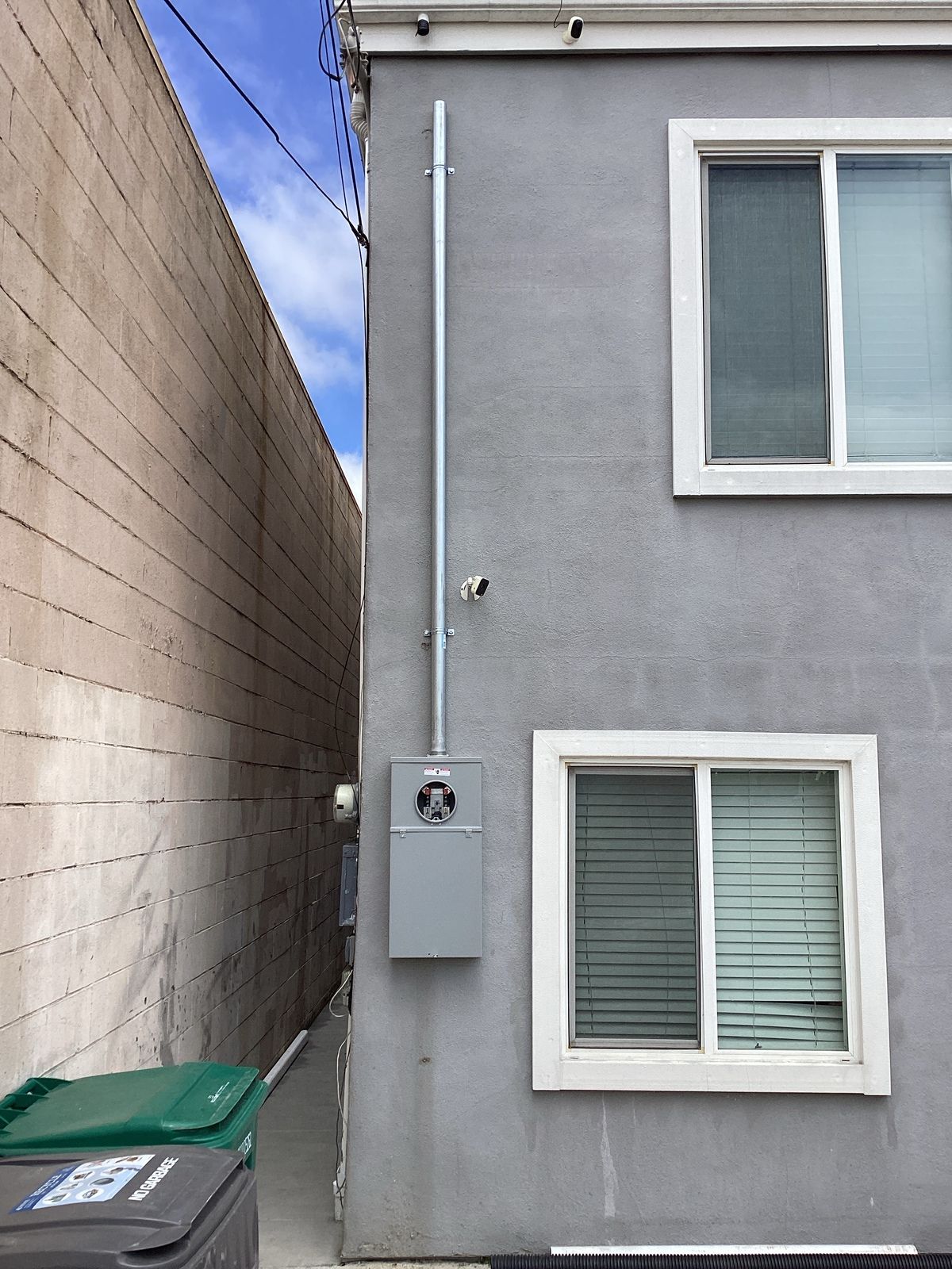 Electrical meter and conduit on a gray building wall, next to a narrow alley. Two windows visible.
