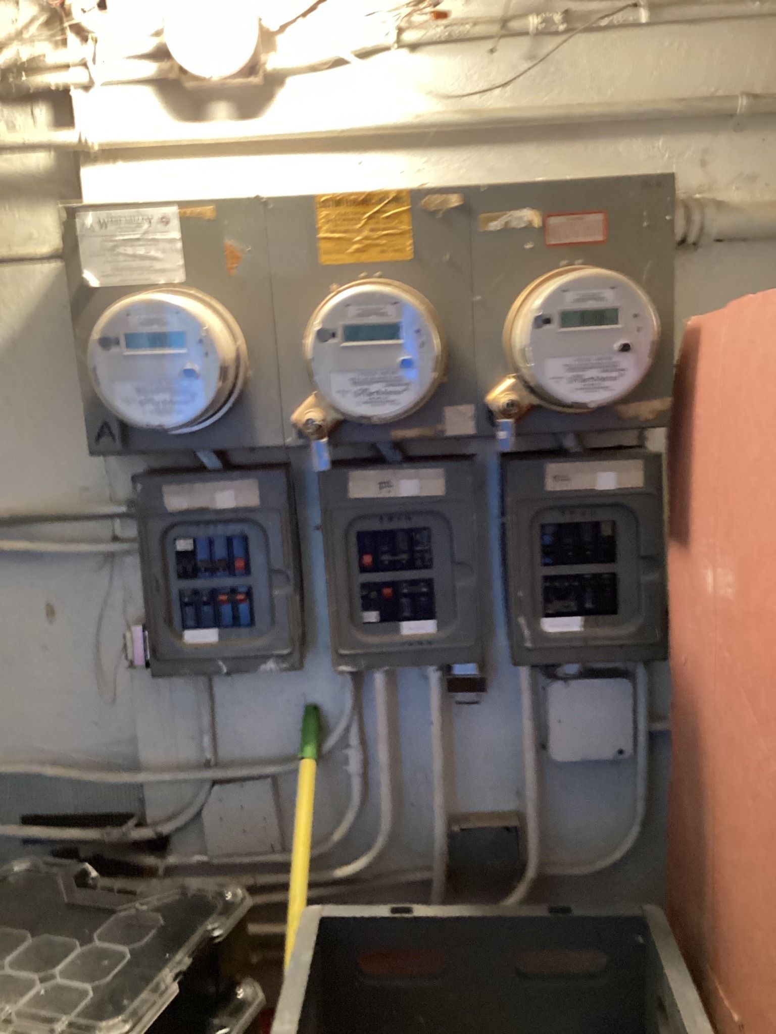 Three electric meters and breaker boxes mounted on a gray wall in a utility room.