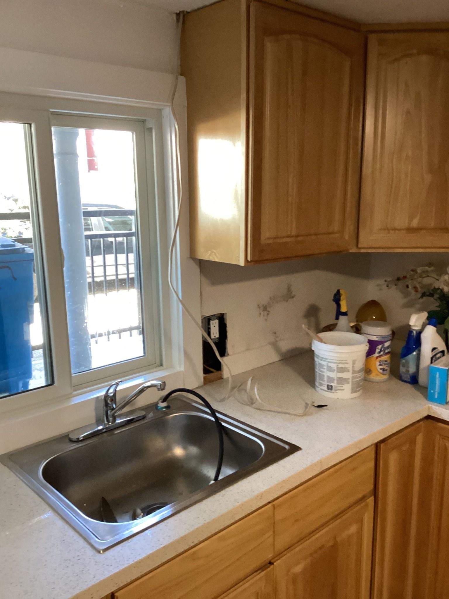Kitchen sink and countertops, cabinets above. Window left, electrical outlet exposed.