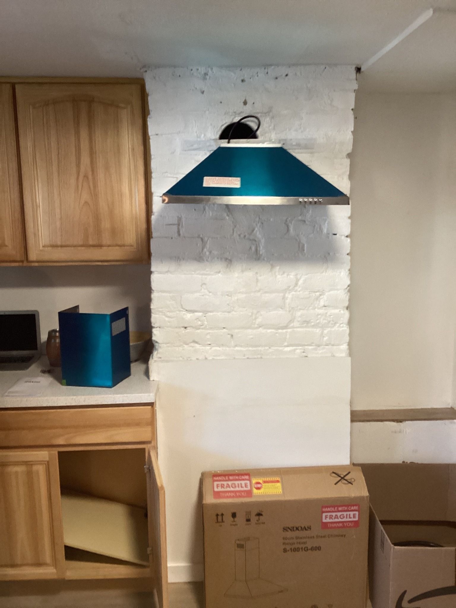 Blue range hood on a painted brick wall in a kitchen with wooden cabinets and a cardboard box.