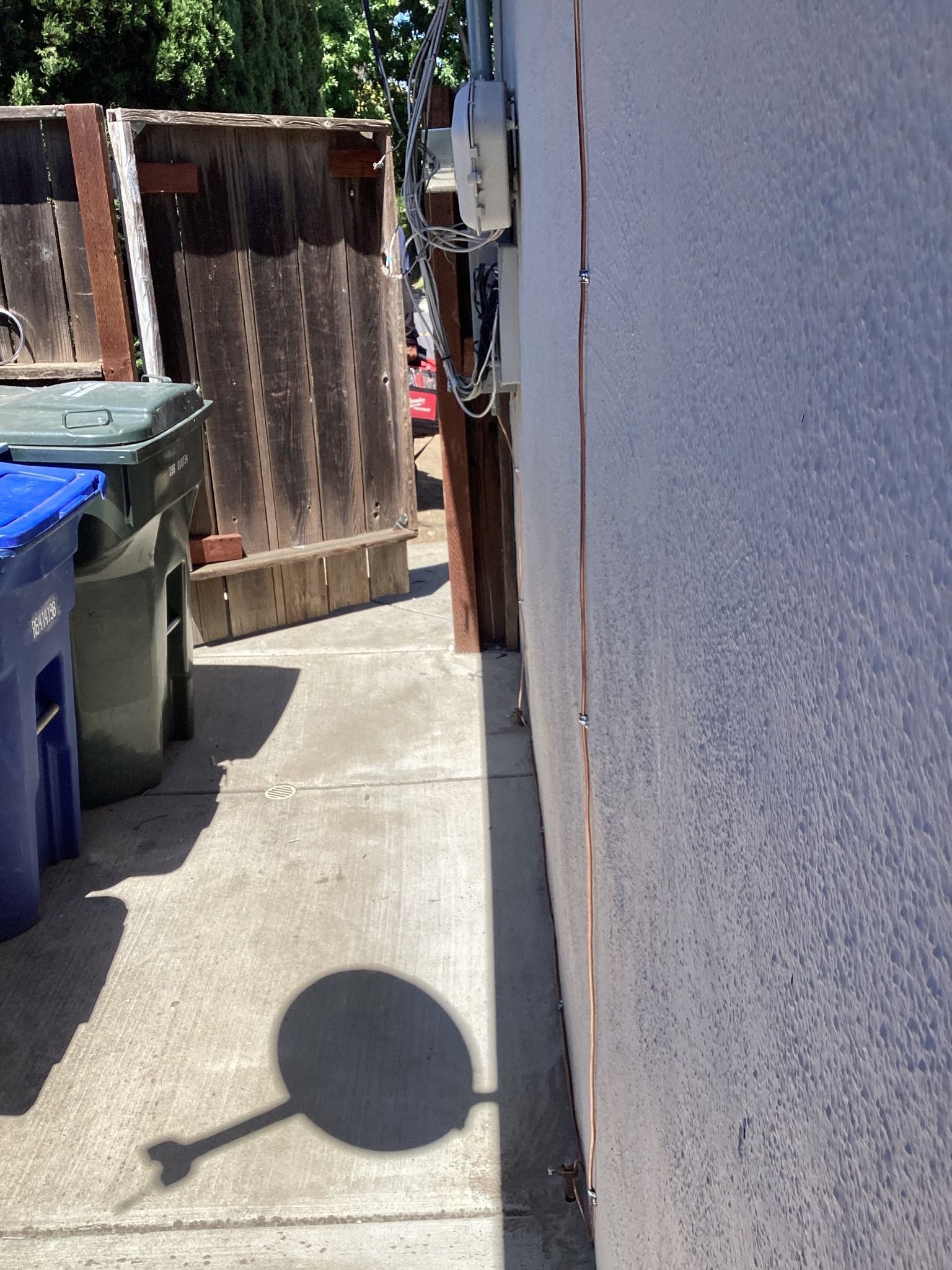 Exterior view of a wall, fence, trash bins, and electrical panel in sunlight. The shadow of a pole is cast on the concrete.