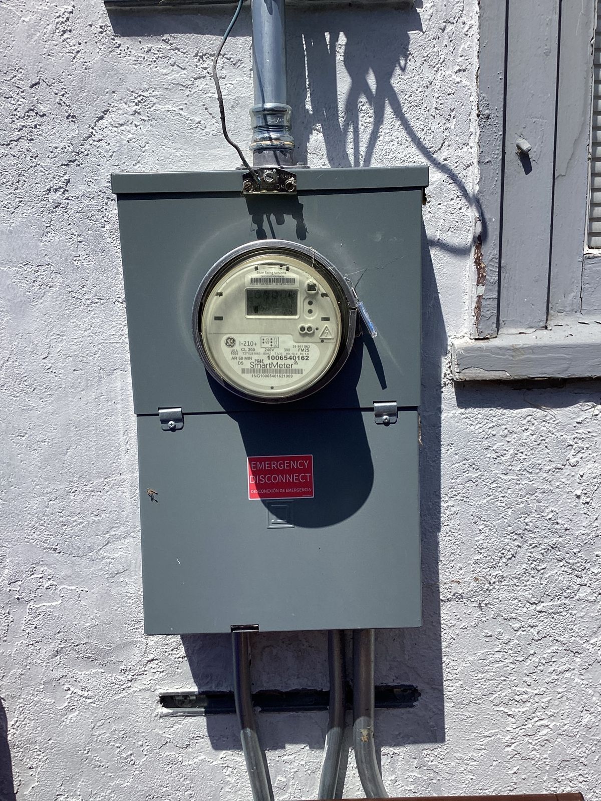 Gray electrical meter box on a stucco wall. Conduit and wires extend from the bottom and top.