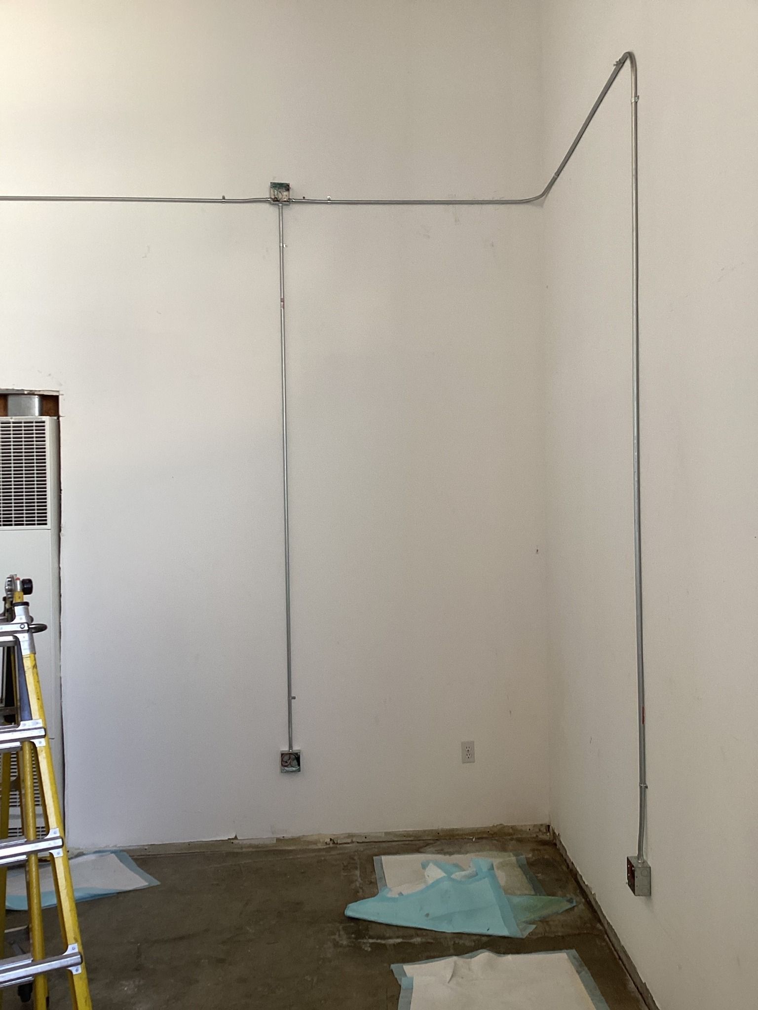 Electrical conduit runs along a white wall, connecting boxes. A ladder sits to the left.