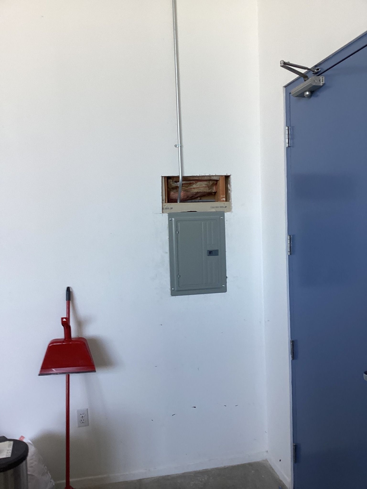 Electrical panel mounted on a white wall; metal conduit overhead; red dustpan on floor.