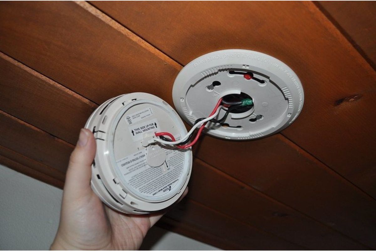 Hand holding a smoke detector, wires attached, near the detector base on a wood ceiling.